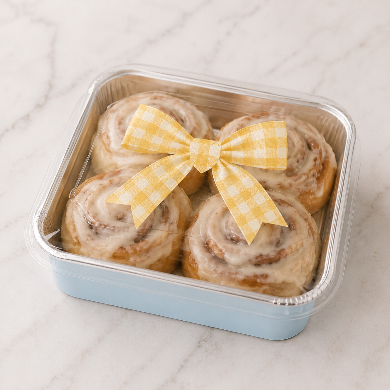 Four glazed cinnamon rolls in a square light blue plastic container with a clear lid, tied with a yellow gingham bow, on a white marble surface.