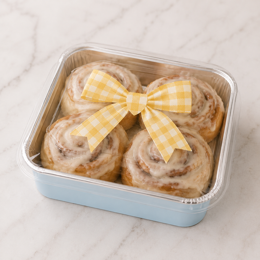 Four glazed cinnamon rolls in a square light blue plastic container with a clear lid, tied with a yellow gingham bow, on a white marble surface.